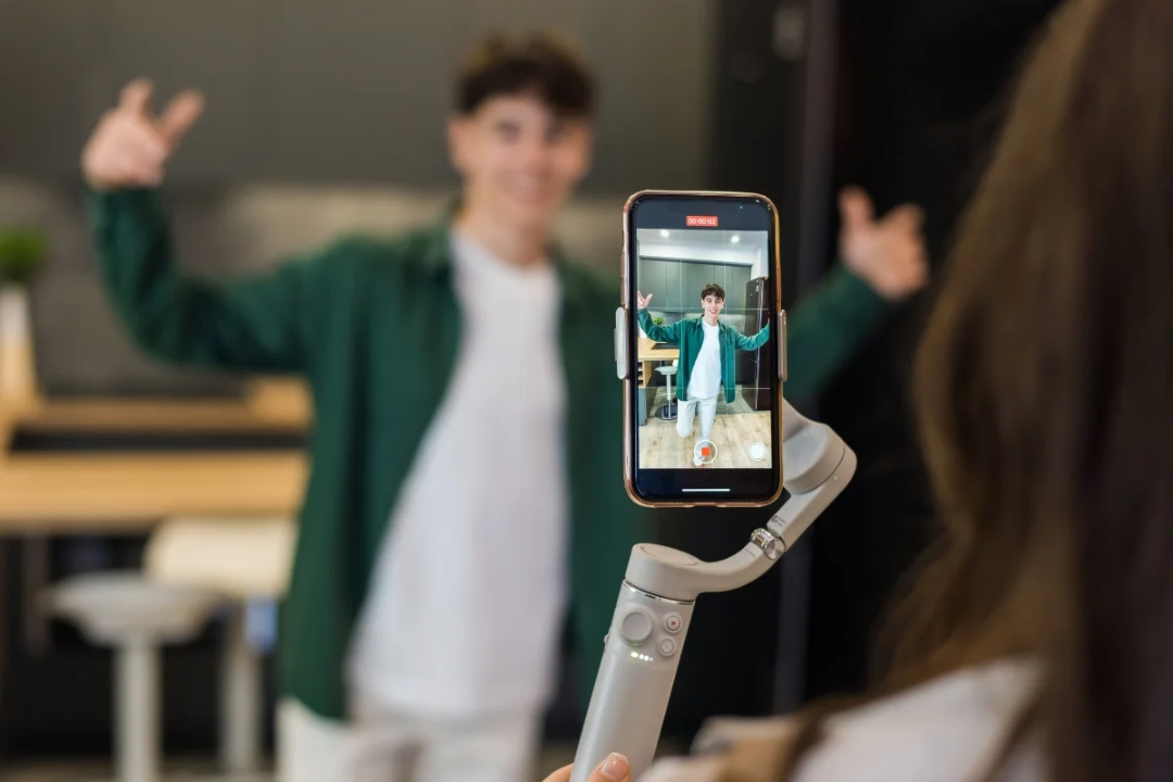 A person records a video of a smiling young man posing with thumbs up using a smartphone mounted on a handheld stabilizer indoors. The phone screen clearly shows the man in focus.