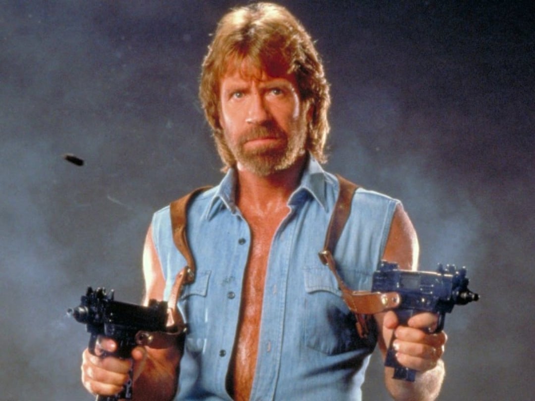 A bearded man with long hair wears a sleeveless denim shirt and leather shoulder holsters, holding two guns and looking intensely forward against a smoky, dramatic background.