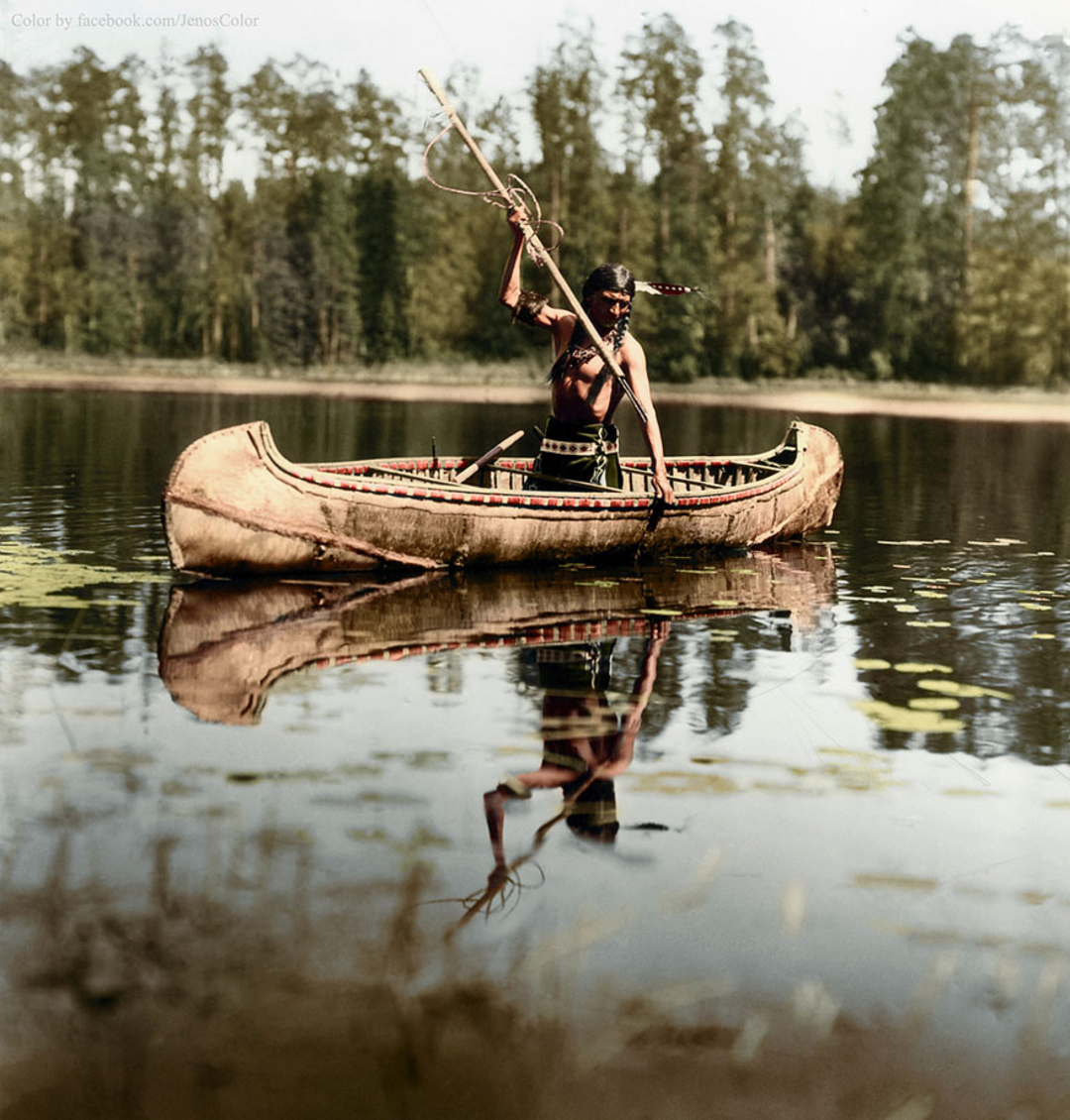 A Native American man in traditional dress paddles a birchbark canoe across a calm lake, surrounded by trees, with his reflection visible in the water.