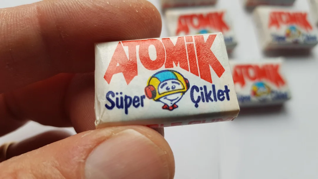 A hand holding a small wrapped gum labeled "ATOMİK Süper Çiklet" with a cartoon smiling face wearing a colorful helmet. Several more gum pieces are blurred in the background.
