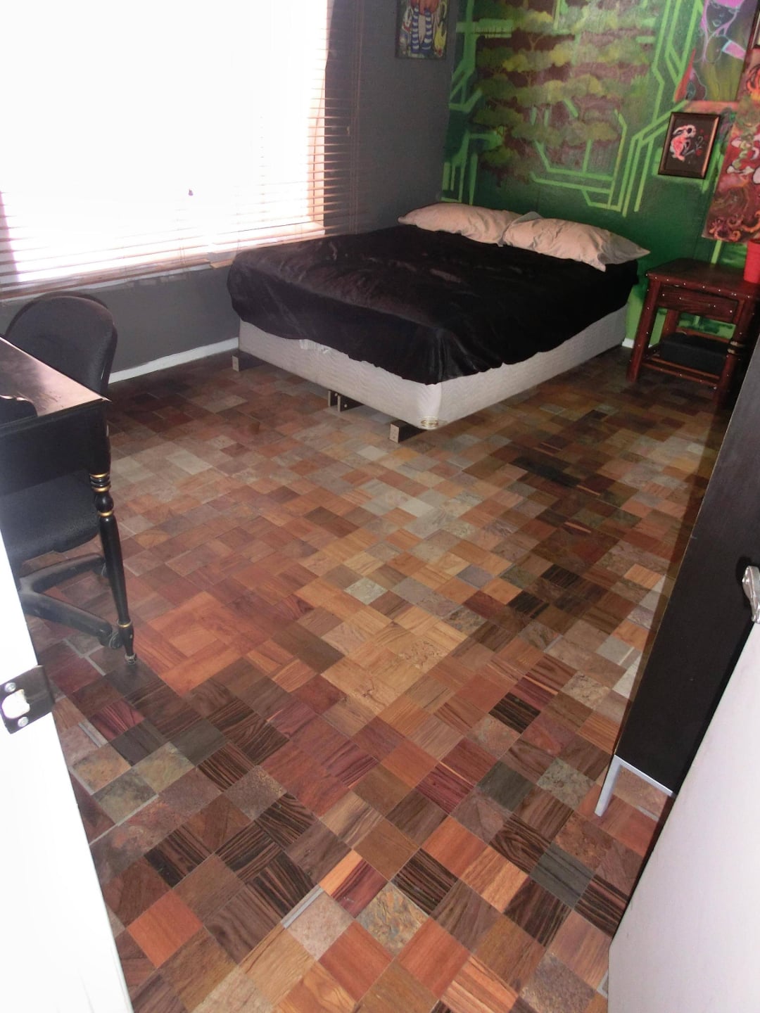 A bedroom with a patchwork-style parquet floor, a bed with black and white bedding, a desk with a chair, and a window with blinds letting in sunlight. The walls feature colorful and abstract artwork.