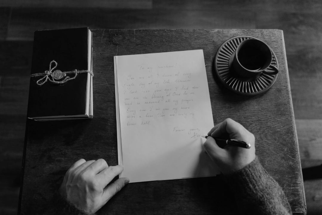 A person writes a letter on white paper at a small table with a notebook tied with string and a cup of coffee on a saucer; the scene is in black and white.