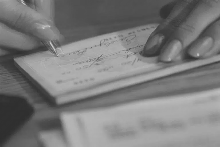 A close-up of a hand holding a pen and writing on a check, with another check or paper partially visible on a wooden surface in the foreground.