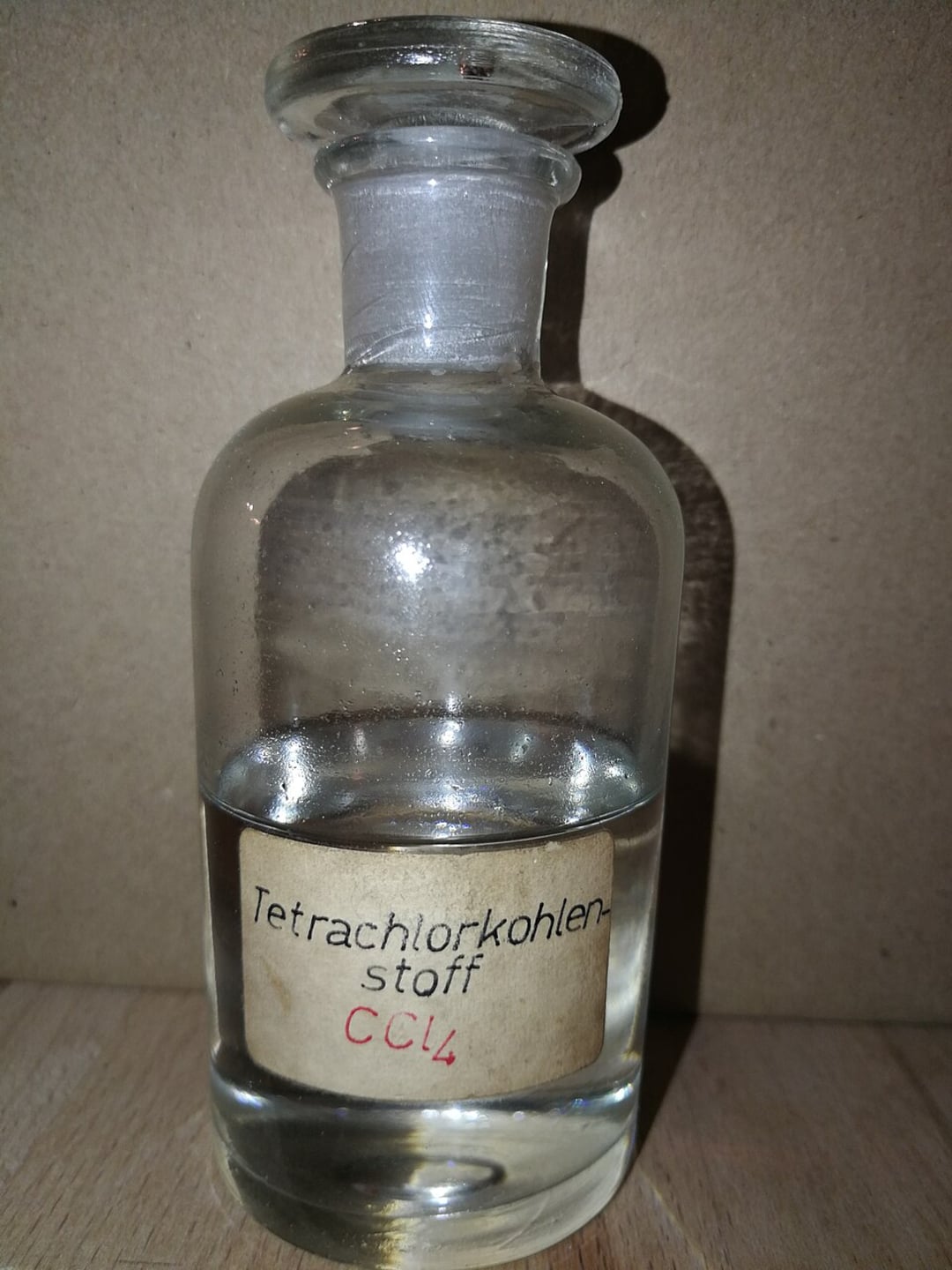 A clear glass bottle with a stopper containing a transparent liquid, labeled "Tetrachlorkohlenstoff CCl4" on a beige paper label, set against a plain brown background.