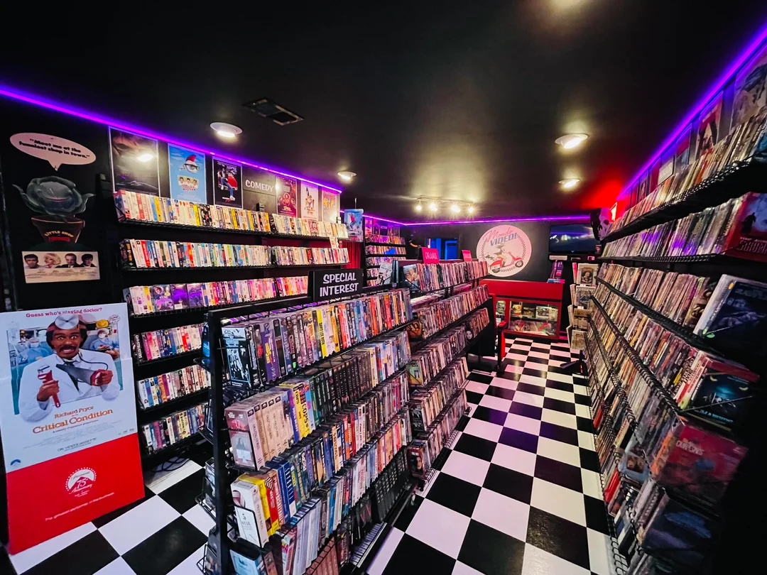A retro video rental store with black-and-white checkered floors, shelves packed with DVDs and VHS tapes, neon purple lights, and a cardboard standee near the entrance. A red and white sign is visible at the back wall.