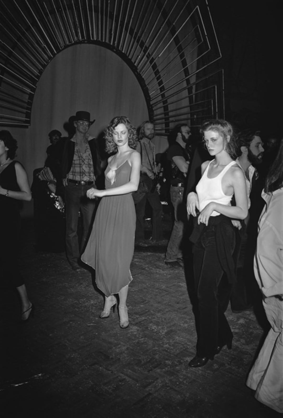 Two women in stylish outfits stand on a dark, crowded dance floor. People mill around behind them, and a large decorative arch frames the scene, creating a lively, retro party atmosphere.