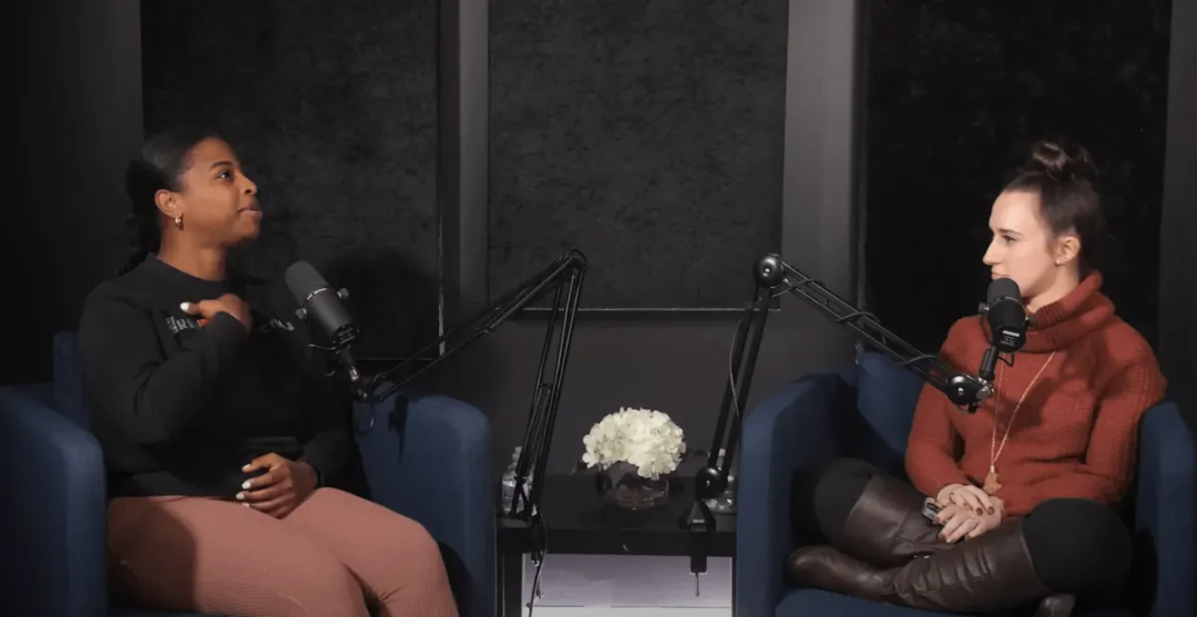 Two women sit in armchairs facing each other, speaking into microphones in a podcast studio. A small table with white flowers is between them, and dark acoustic panels are on the wall behind them.