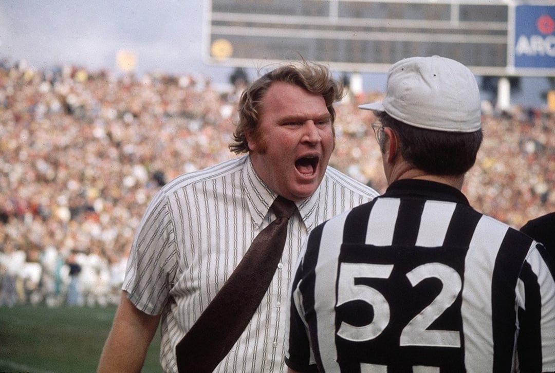 A football coach in a striped shirt and brown tie is yelling at a referee wearing a black-and-white striped shirt with the number 52 on the back. A packed stadium is visible in the background.