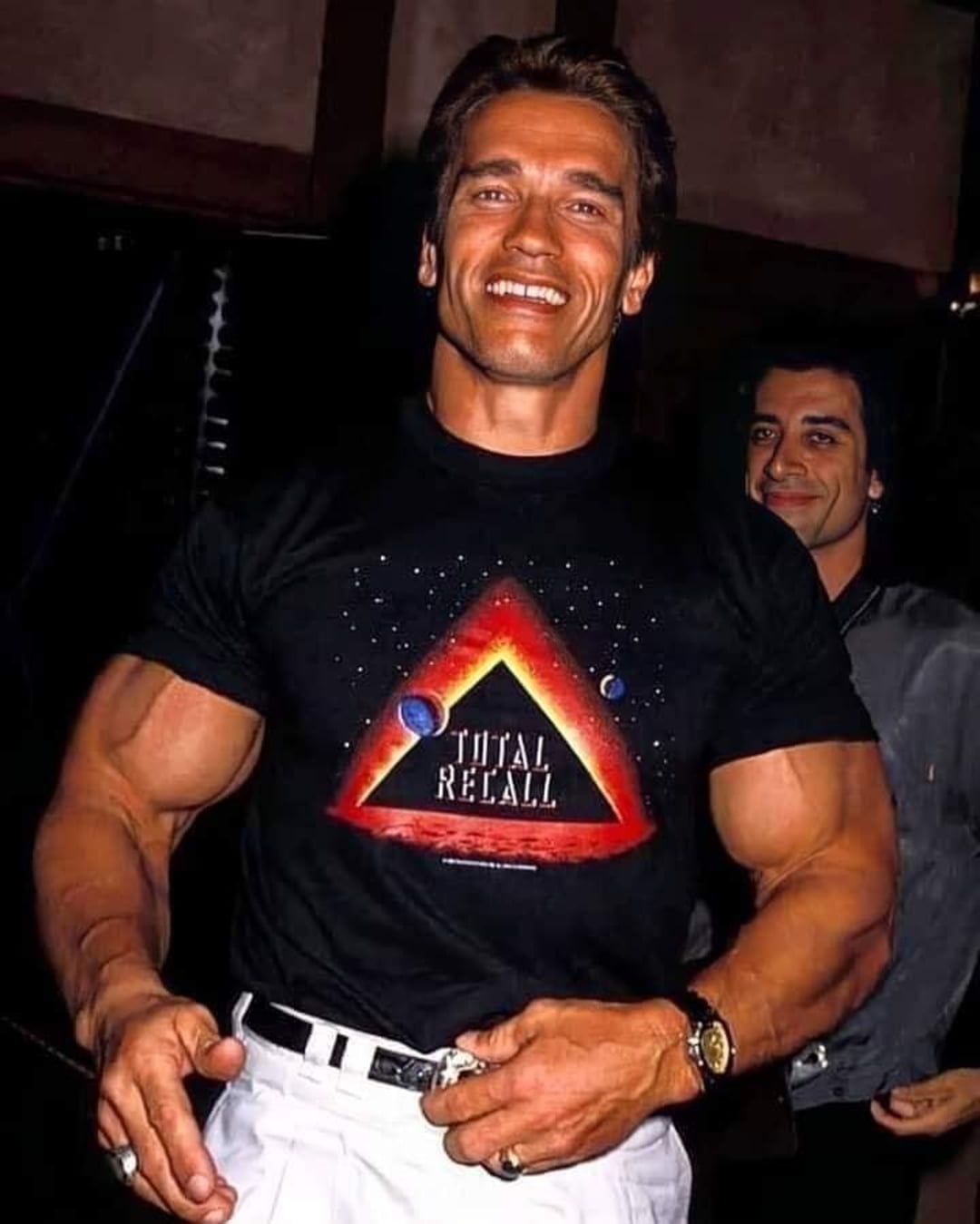 A muscular man smiling, wearing a black "Total Recall" T-shirt with a triangular logo and white pants. Another man in the background is also smiling.