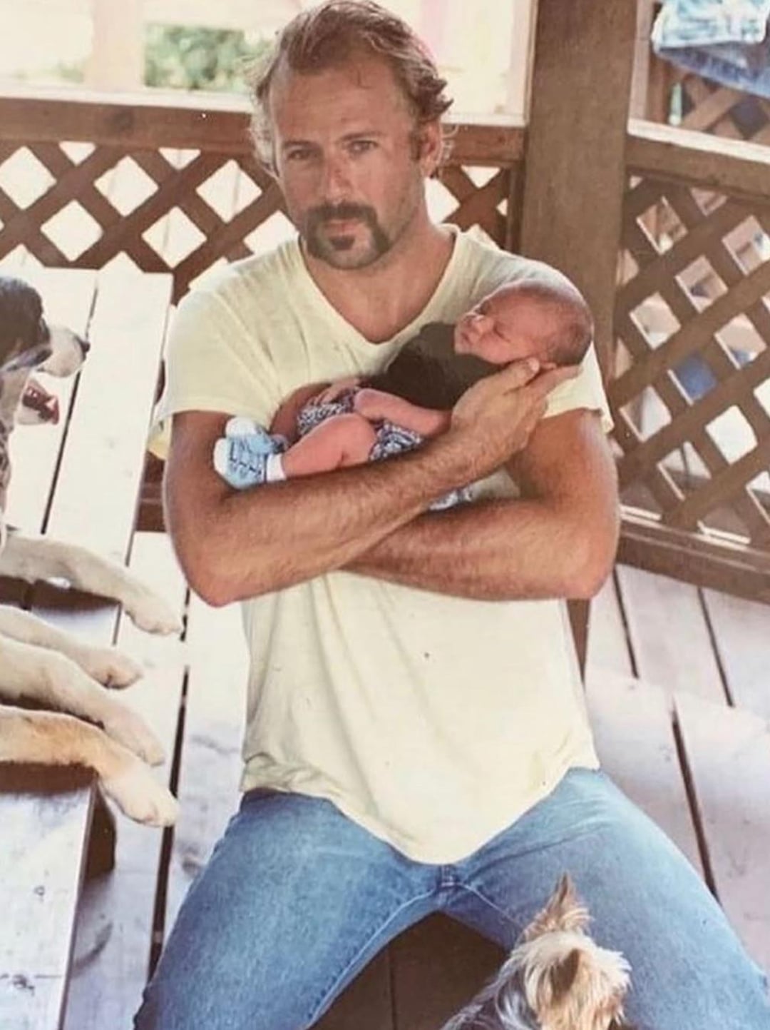 A man with light hair and a mustache sits on a wooden porch, holding a newborn baby in his arms. He wears a light t-shirt and jeans. Several dogs are partially visible around him.