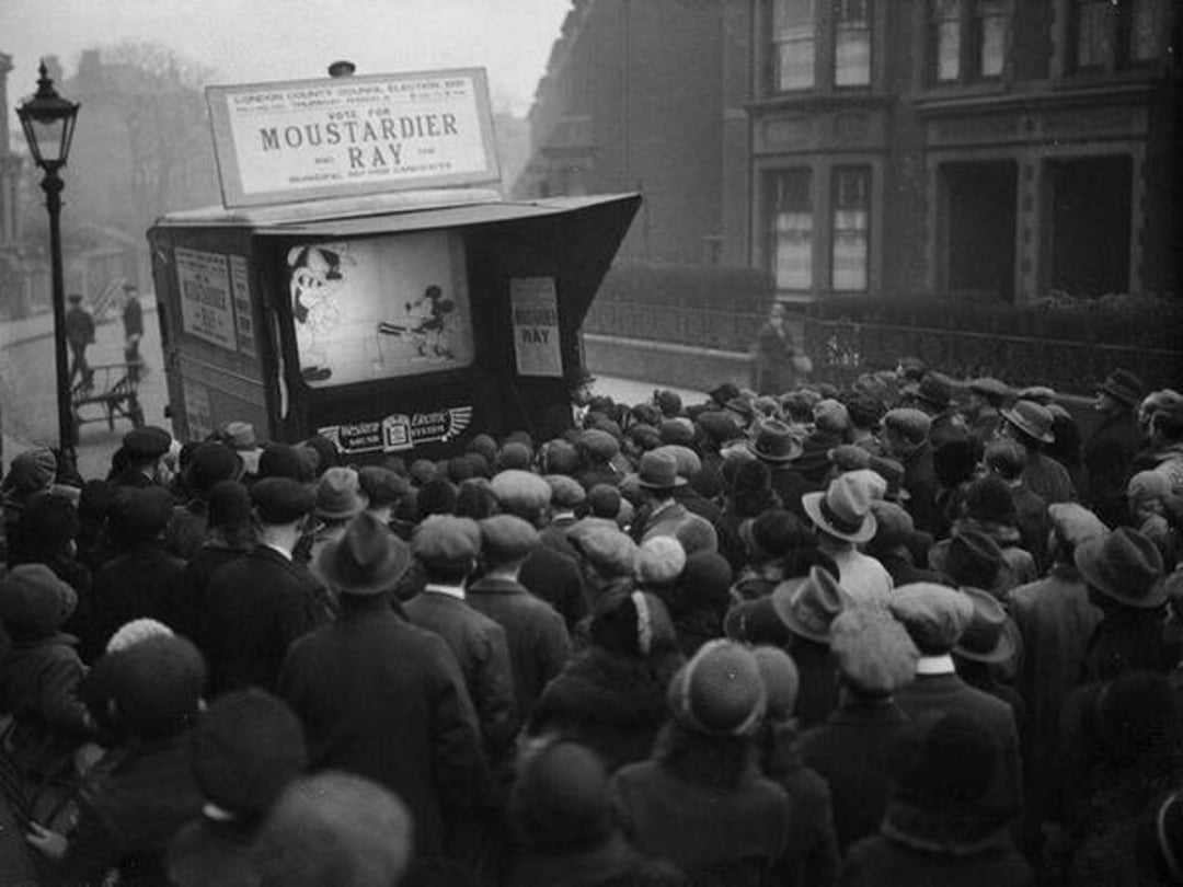 A large crowd of people in hats gathers on a street, watching a cartoon projected from the back of a van labeled "Moustardier Ray." The scene appears to be from the early 20th century.