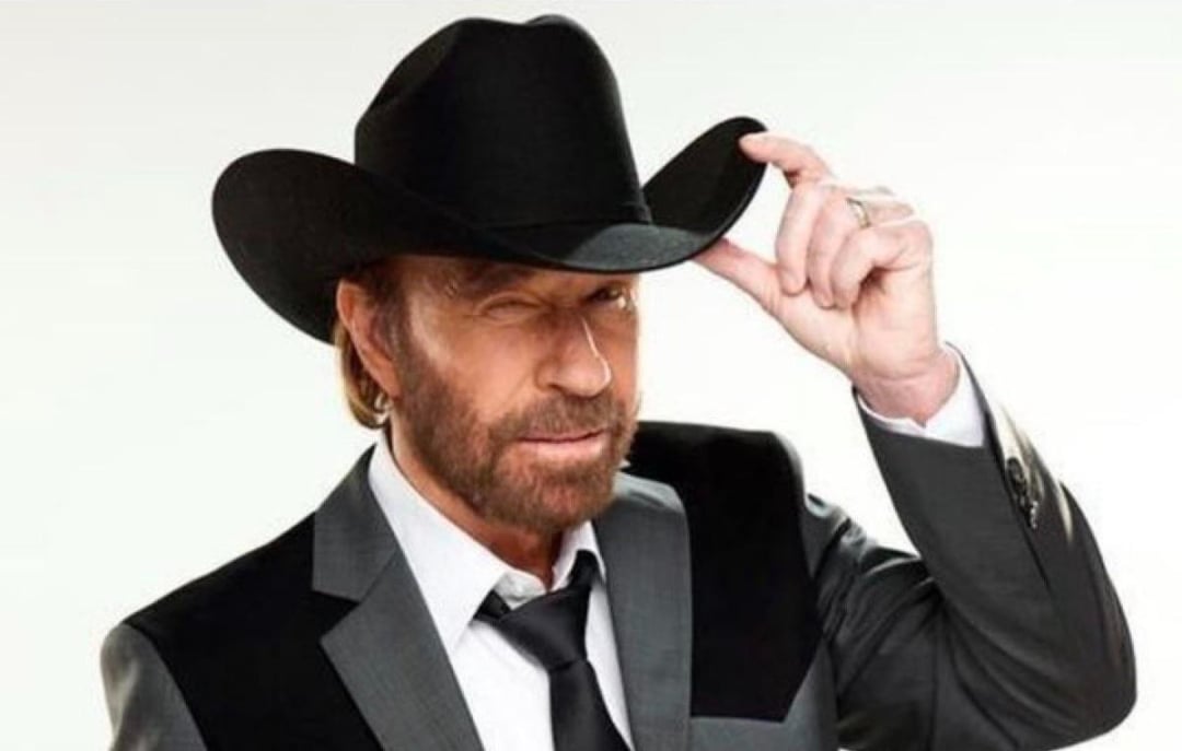 A man with a beard wearing a black cowboy hat and a gray suit with a black tie, tilts his hat and winks at the camera against a plain light background.