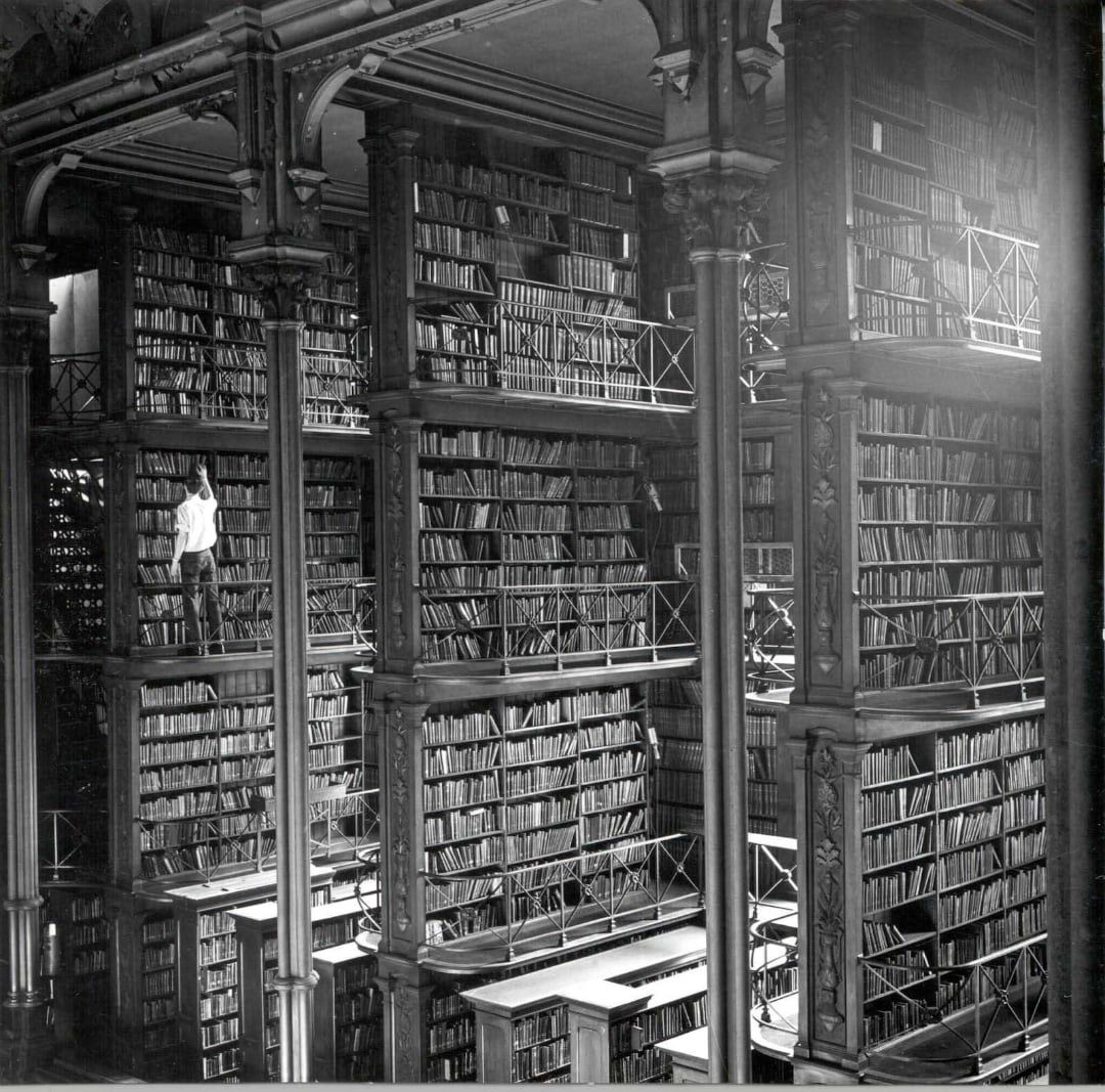 A person stands on a narrow balcony among tall, multi-level bookshelves filled with books in a grand, ornate library with high ceilings and large pillars.