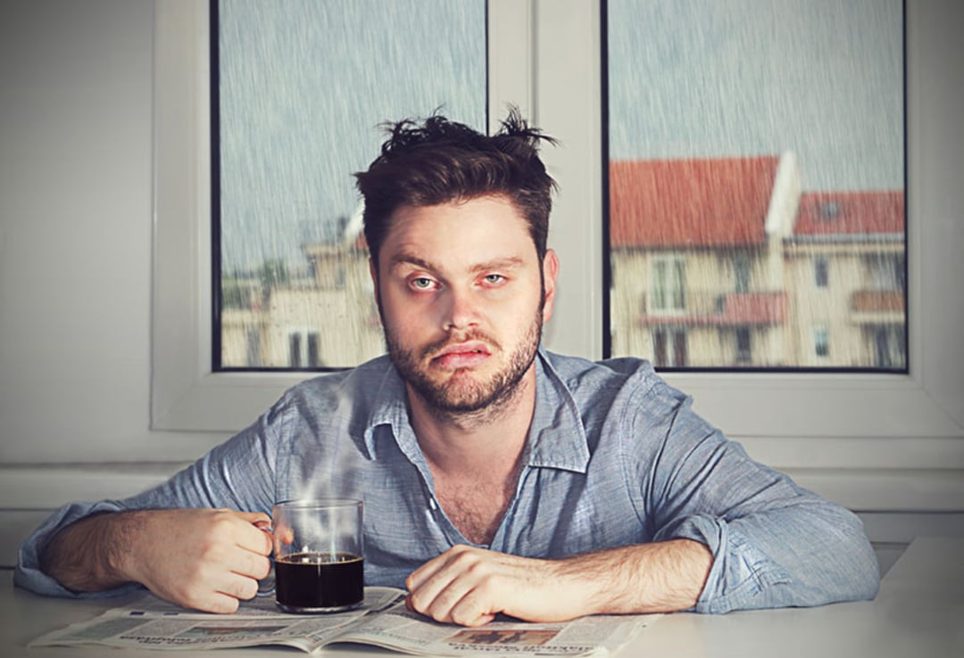 A tired man with messy hair and a scruffy beard sits at a table holding a cup of coffee, looking exhausted. There’s a newspaper on the table and a rainy window with blurred buildings in the background.
