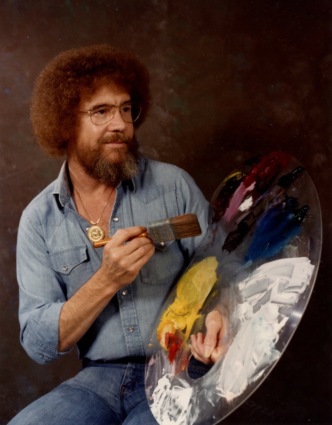 A man with curly hair, glasses, and a beard, wearing a denim shirt, holds a large paint palette and brush, posed as if painting. The background is dark and neutral.