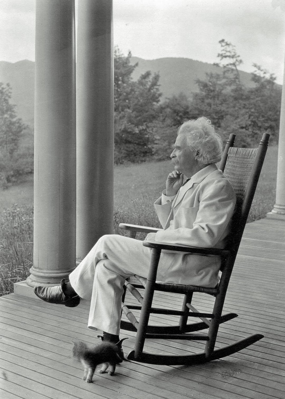 An older man with white hair, dressed in a light-colored suit, sits in a rocking chair on a porch, gazing thoughtfully into the distance. Trees, hills, and a small dog are visible in the background.
