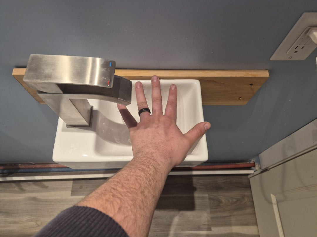 A person's hand, wearing a black ring, is shown above a small white sink with a modern faucet mounted on a wooden board against a gray wall. The hand appears too large for the compact sink.
