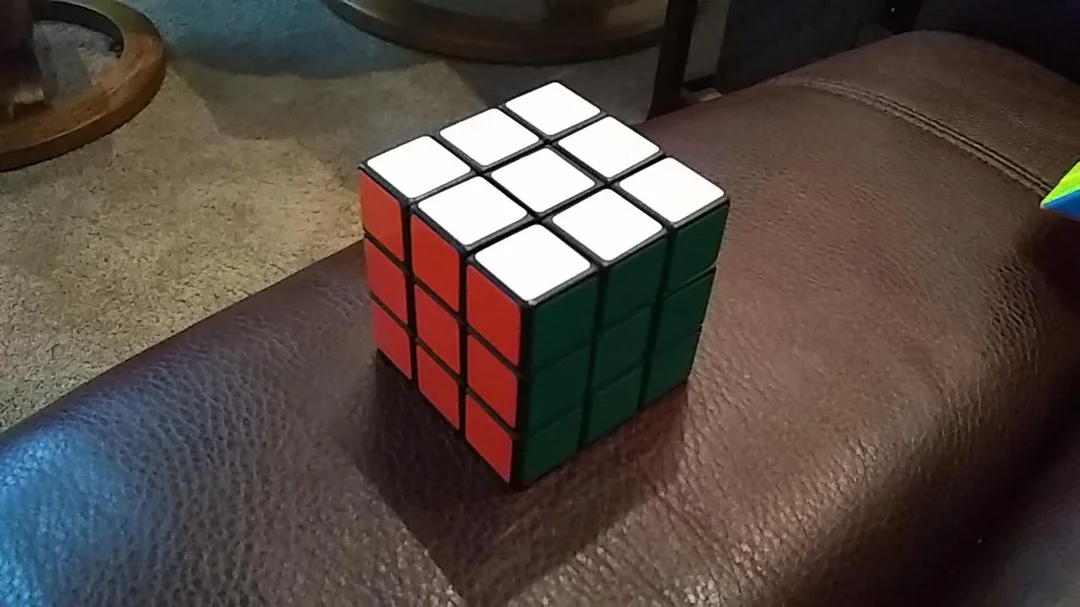 A solved Rubik's Cube is resting on a brown leather surface, showing white, red, and green faces. Carpet and furniture are visible in the background.