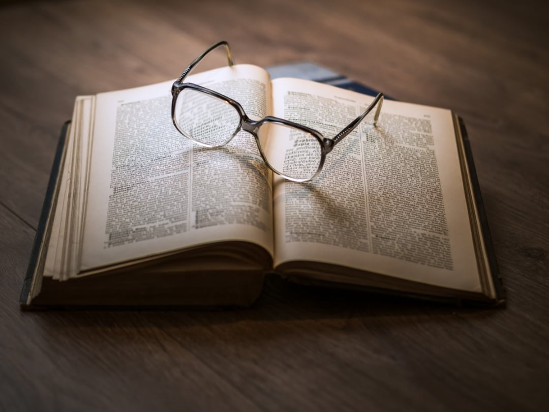 A pair of eyeglasses rests on the open pages of a thick book lying on a wooden surface, with light softly illuminating the text.