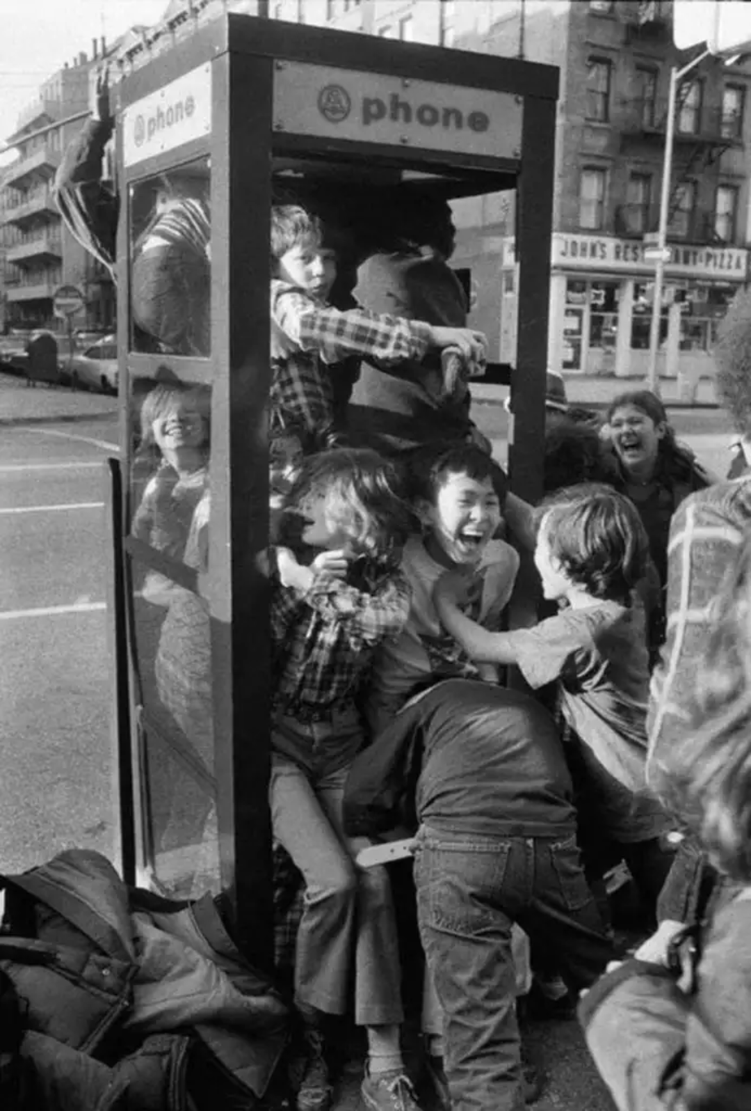 A group of smiling and laughing children squeeze inside and around a crowded phone booth on a city street, playing together joyfully.