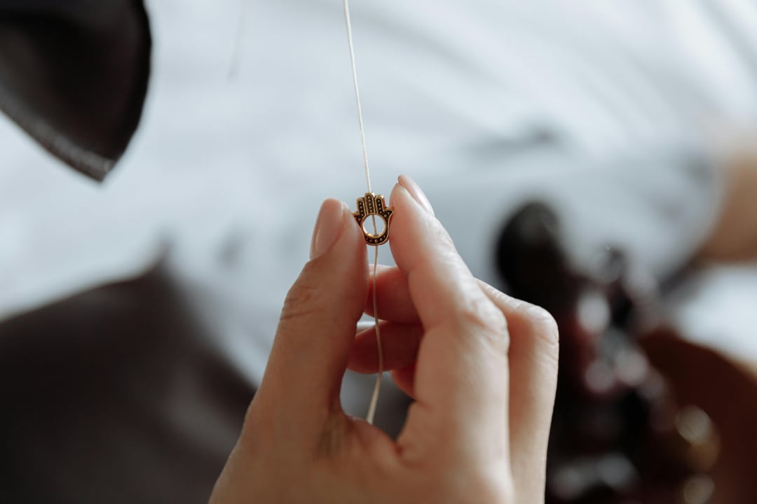 A close-up of a hand holding a string with a small gold Hamsa hand pendant, against a blurred background.