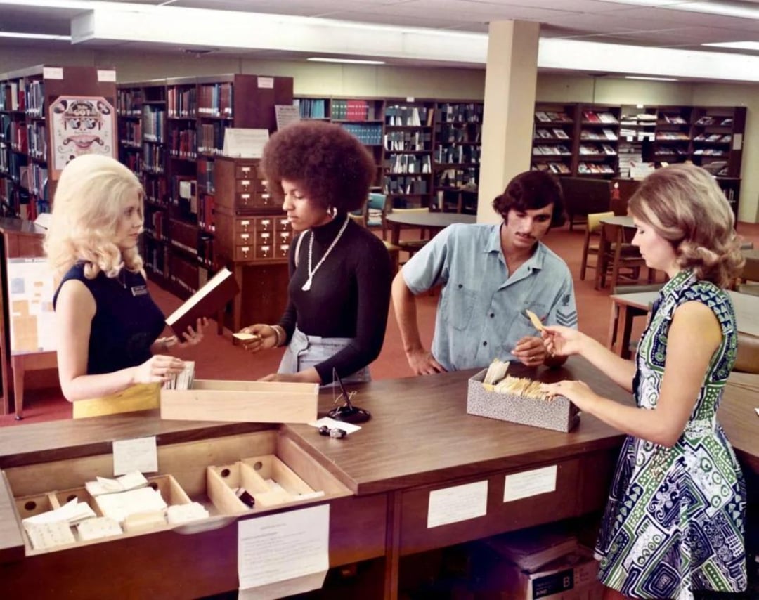 Four people, three women and one man, stand at a library counter with card catalogs and books. Bookshelves filled with books are visible in the background. The scene appears to be from the 1970s.