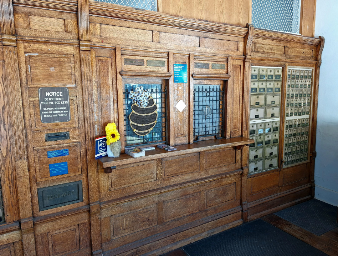 An old-fashioned wooden post office counter with small mailboxes, a bulletin board, a vase with yellow flowers, a beehive decoration, and various signs and notices on the wall.