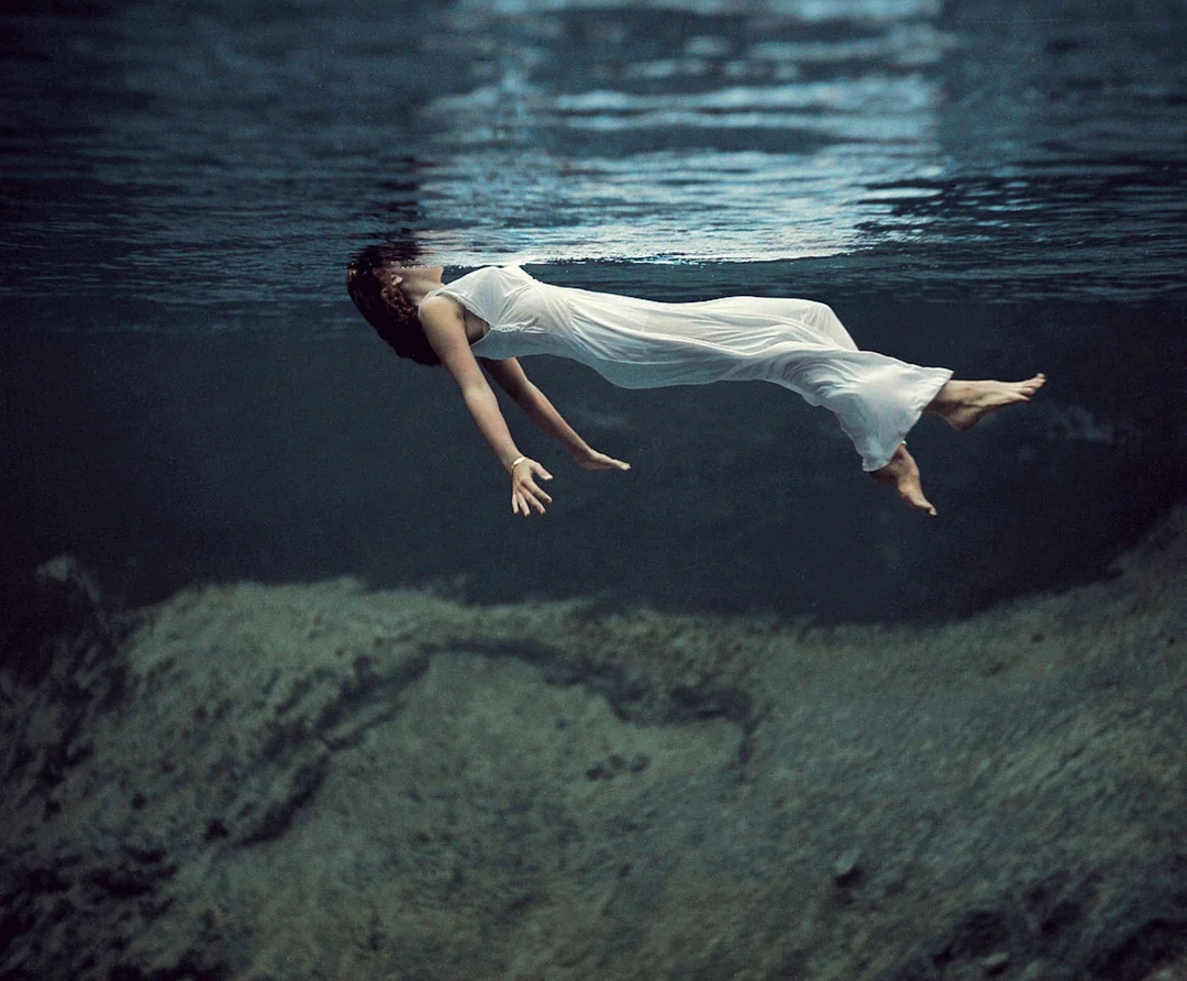 A woman in a white dress floats serenely underwater, her body parallel to the surface and her dark hair flowing around her, with a rocky bottom visible below.