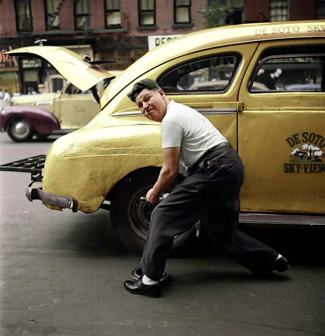 A man in a white shirt and dark pants changes the rear tire of a yellow De Soto taxi on a city street, with the car trunk open and old buildings visible in the background.