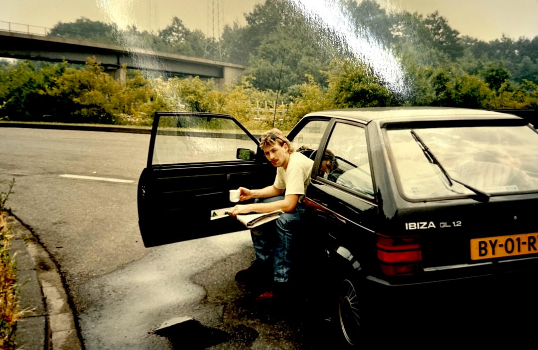 A man sits in the open driver’s door of a black SEAT Ibiza car parked by the roadside, holding a cup and looking at the camera. Trees, bushes, and an overpass are visible in the background.