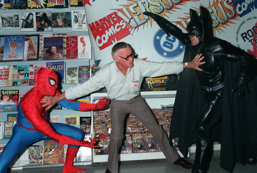 Stan Lee playfully poses between people dressed as Spider-Man and Batman at a comic book store, separating them with his arms. Comic books and posters line the walls behind them.