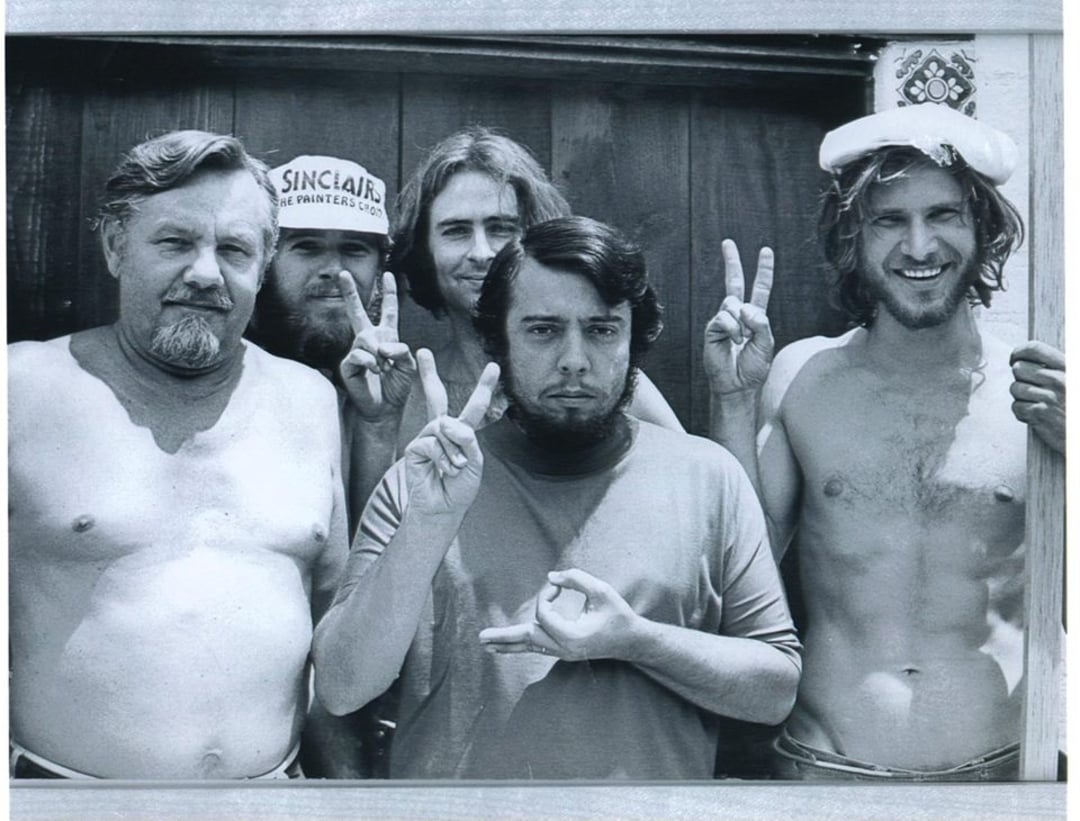 Five men pose for a black-and-white photo, some shirtless, all making peace signs. One man wears a cap, another has a towel or hat on his head; they stand close together and smile or make playful faces.