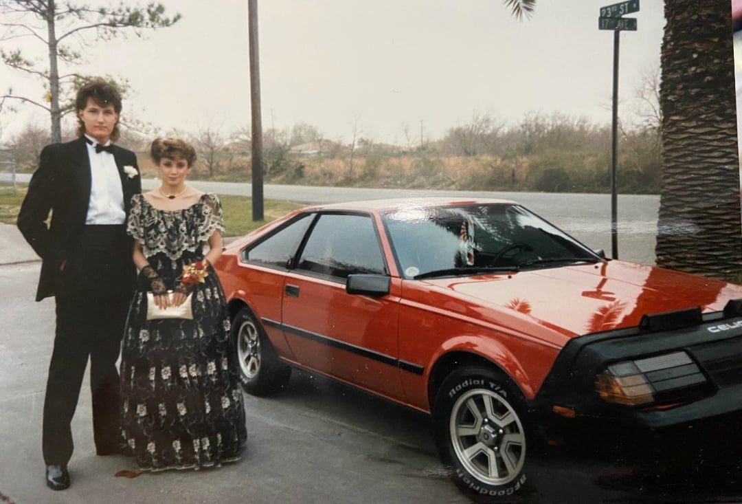 A young man in a tuxedo and a young woman in a formal dress stand beside a red sports car on a street, with trees and a street sign visible in the background.