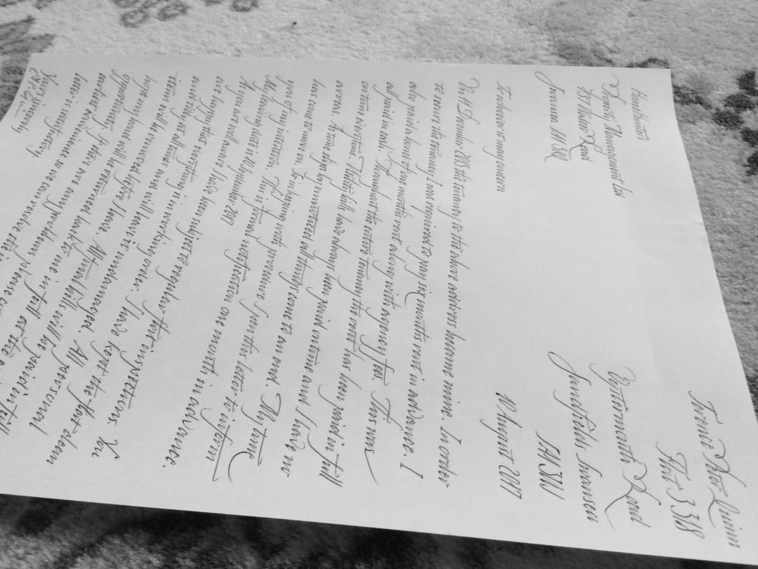 A black and white photo of a handwritten letter with elegant cursive script, placed on a textured surface. The letter appears formal and decorative, with flourishes and structured lines of text.