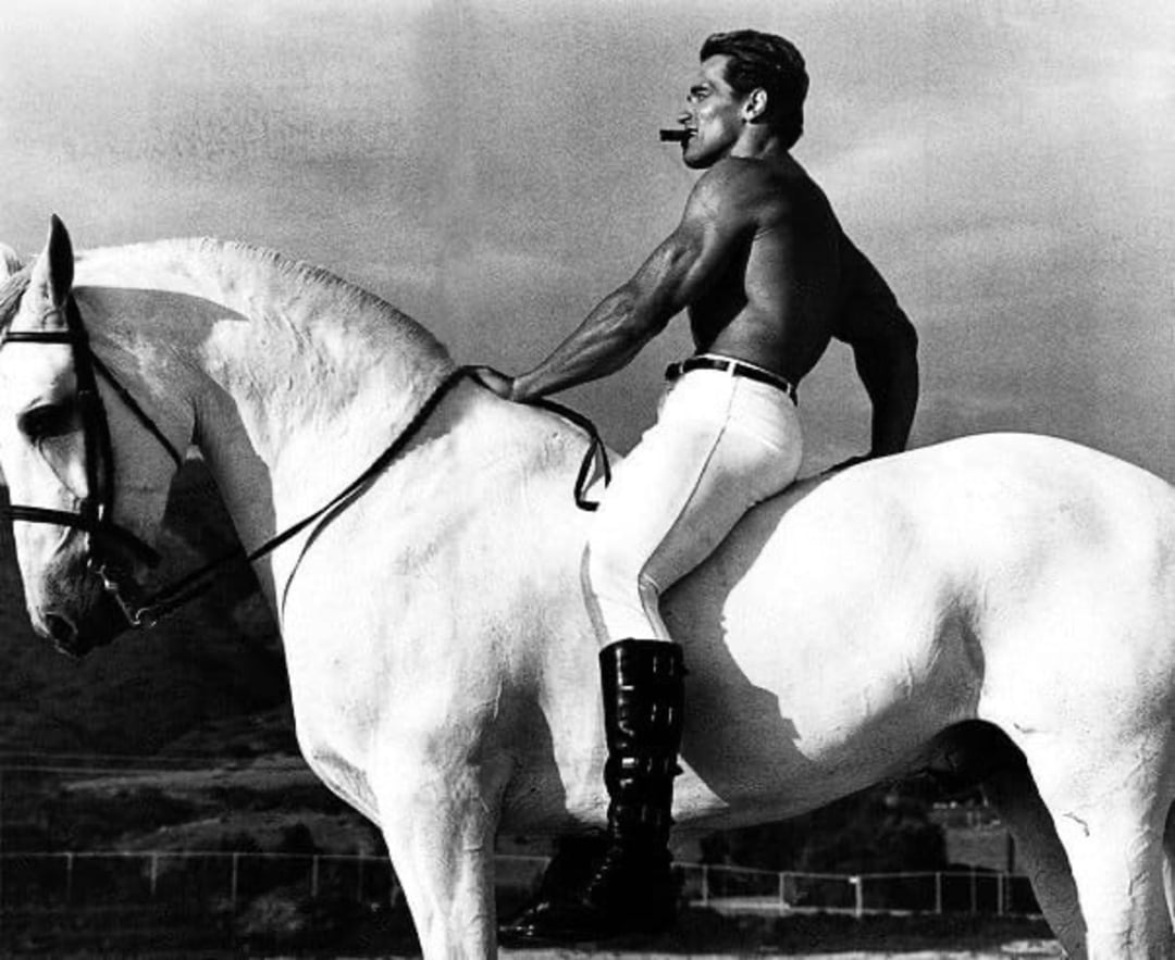 A muscular, shirtless man wearing white pants and black boots sits confidently on a large white horse, holding the reins with one hand and smoking a cigar. The background appears to be an outdoor setting.