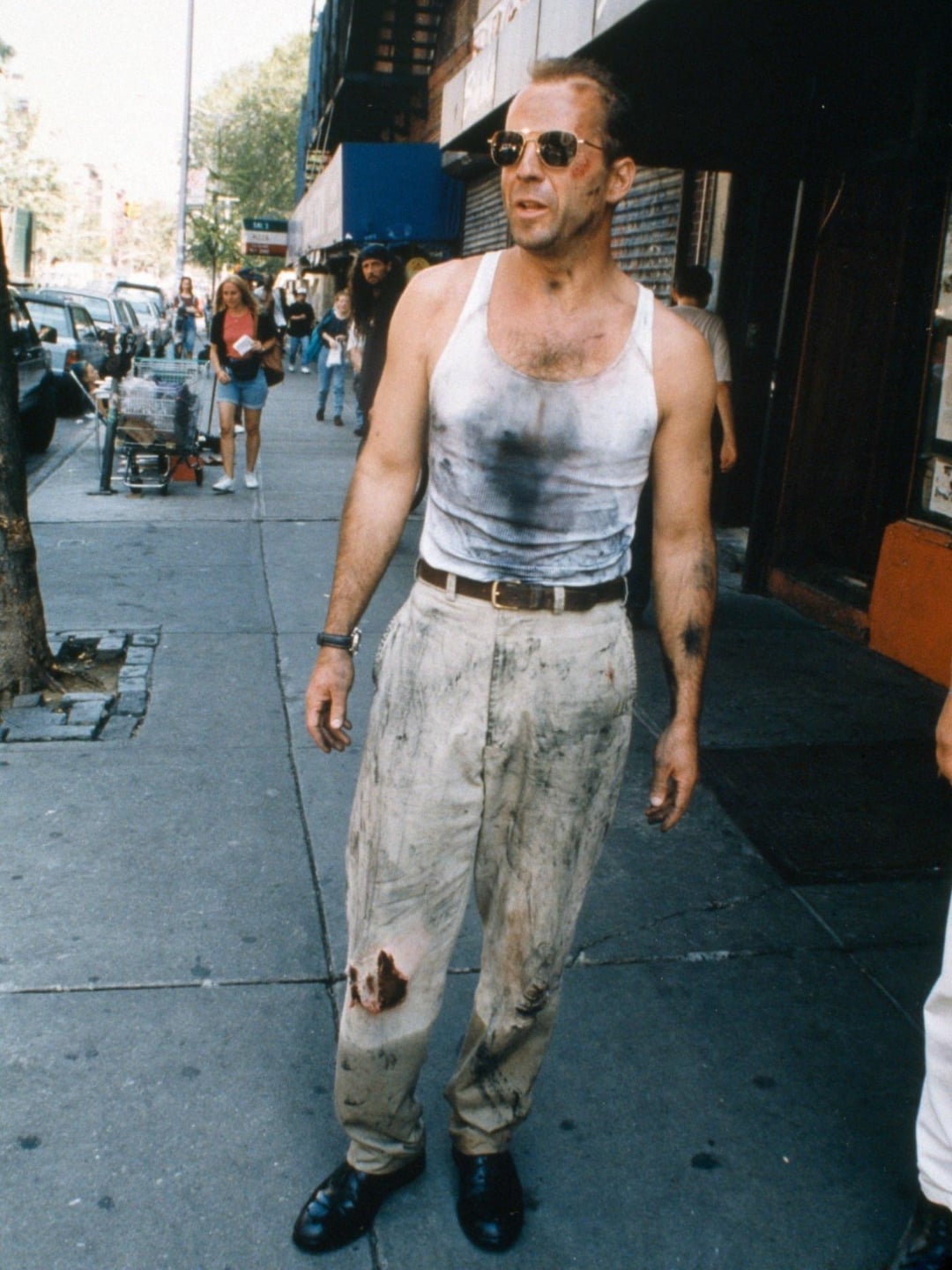 A man in a dirty white tank top and stained beige pants stands on a city sidewalk, looking to the side. He wears sunglasses and has messy hair, with dirt and grime on his clothes and skin. Urban street scene in the background.