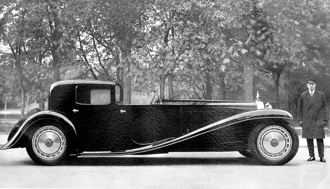 A vintage black luxury car with elegant curved fenders is parked on a tree-lined road. A man in a long coat and hat stands beside it. The photo is in black and white.