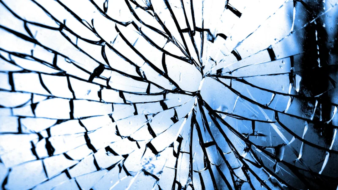 Close-up of shattered glass with sharp cracks radiating outward from the center, creating an intricate web-like pattern. The image is tinted blue, enhancing the dramatic effect of the broken fragments.