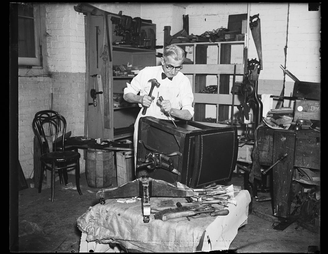 An older man wearing glasses works on upholstering a chair with a hammer and nail in a workshop filled with tools, shelves, and furniture parts.