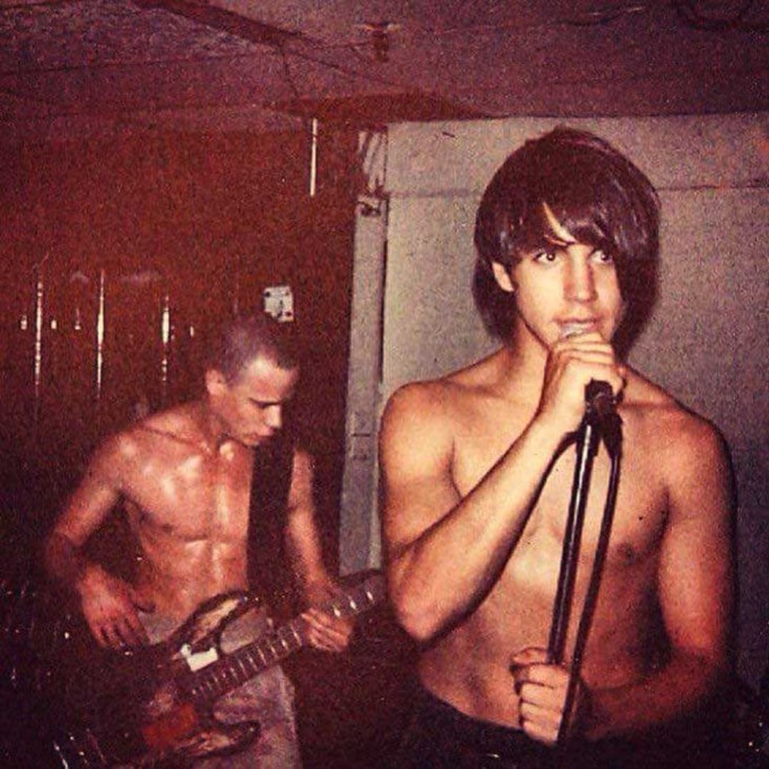 Two young, shirtless men perform in a dimly lit room; one sings into a microphone while the other plays an electric bass guitar in the background.