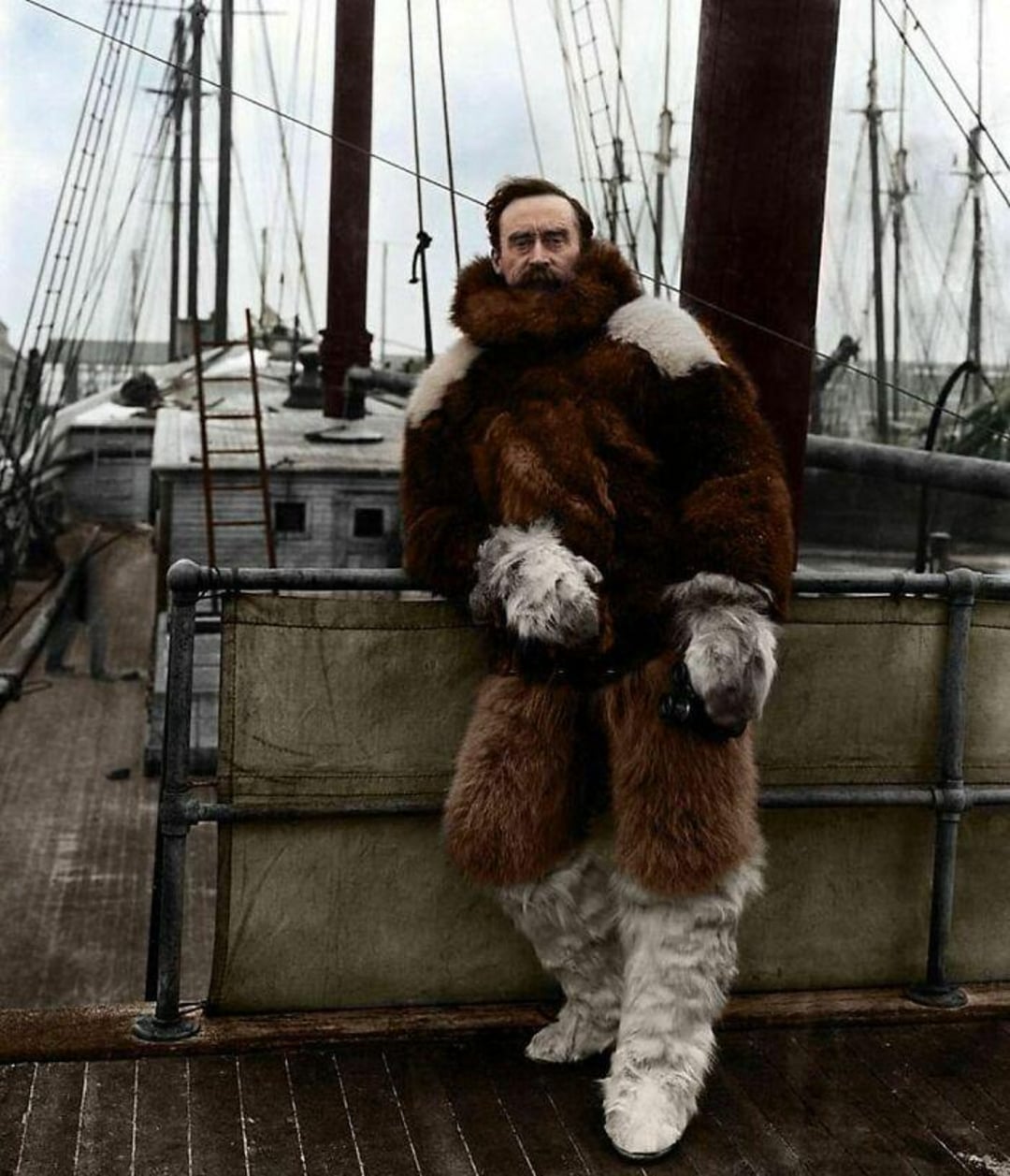 A man in a thick fur coat and boots stands on the deck of a wooden ship, surrounded by masts and rigging. The image appears to be historical, with muted colors and a wintry, expedition-like atmosphere.