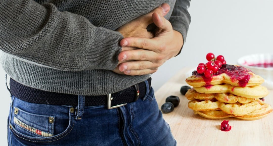 A person in a gray sweater and jeans holds their stomach in apparent discomfort, standing next to a stack of pancakes topped with berries and sauce on a wooden table.
