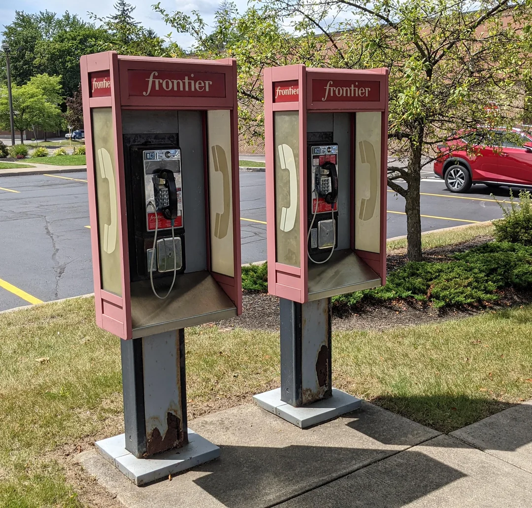 Two old-fashioned Frontier payphones stand side by side on a sidewalk near a parking lot, with some rust on their bases. There are trees, grass, and a red car in the background.