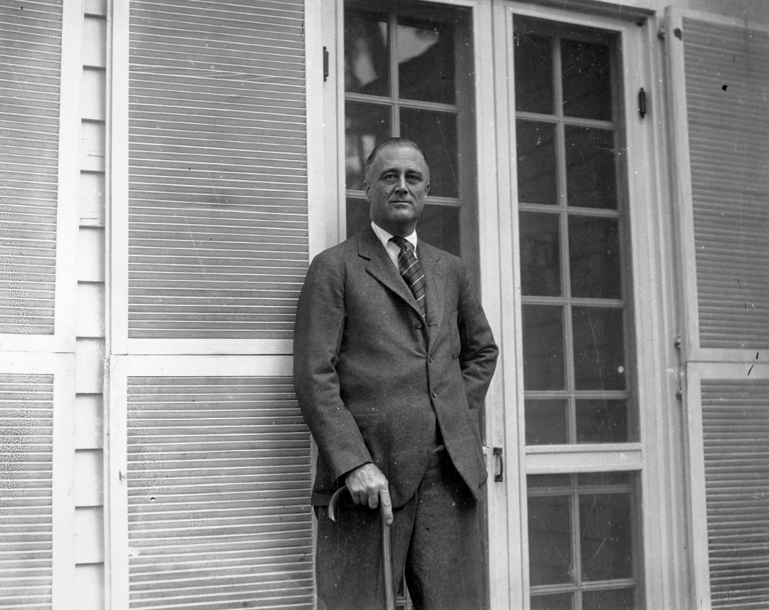 A man in a suit stands outside in front of a building with glass doors and shuttered windows, one hand in his pocket and the other holding a cane.