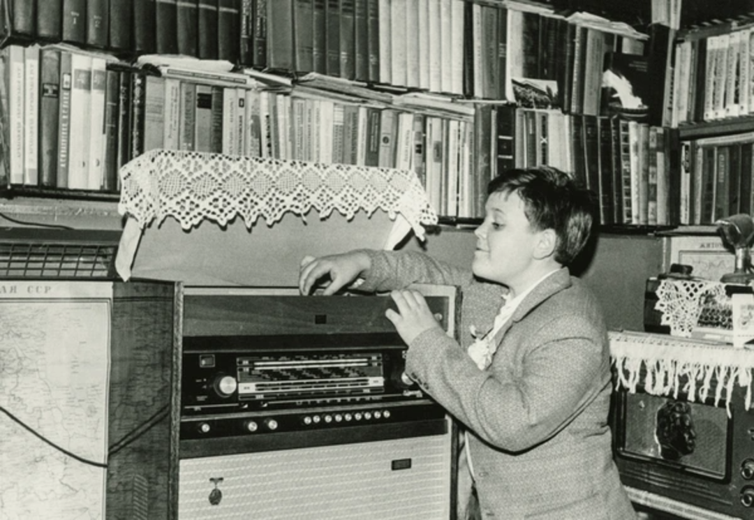 A young boy in a suit adjusts a vintage radio or record player in a room filled with bookshelves, lace doilies, and a map on the wall. The setting appears to be from the mid-20th century.