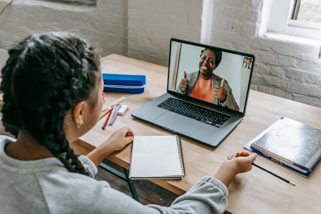A person sits at a desk with notebooks, having a video call on a laptop with someone smiling and giving two thumbs up. The desk has pens and a window is in the background.