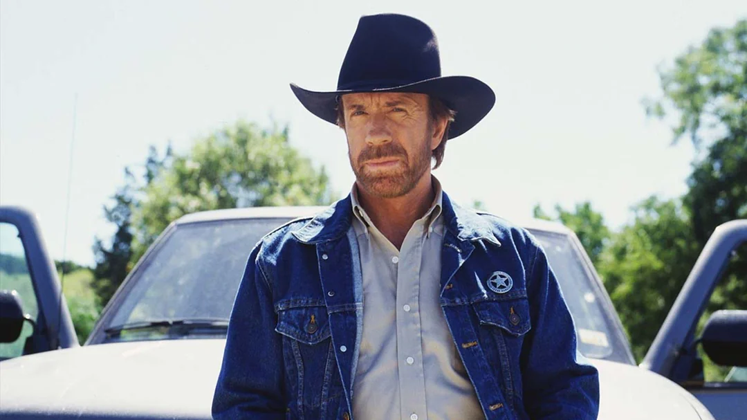 A man wearing a black cowboy hat and denim jacket with a sheriff’s badge stands in front of a white vehicle outdoors, with trees and a blue sky in the background.