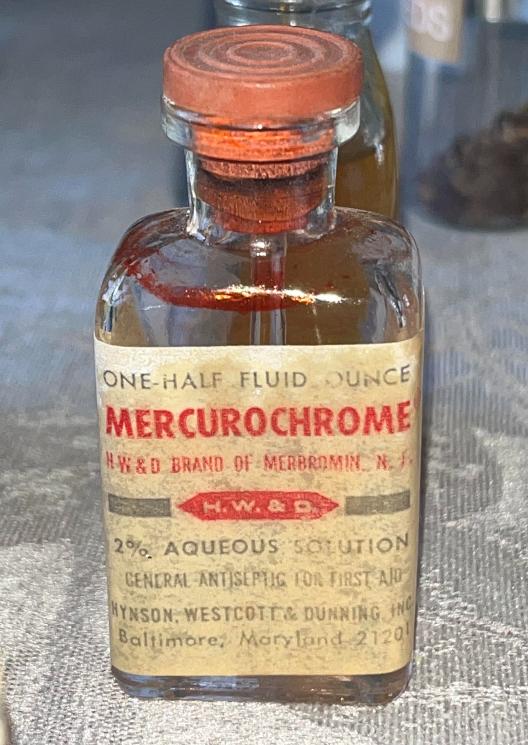 A small vintage glass bottle with a red-orange cap labeled "Mercurochrome," containing a 2% aqueous antiseptic solution for first aid, produced by Hynson, Westcott & Dunning in Baltimore, Maryland.