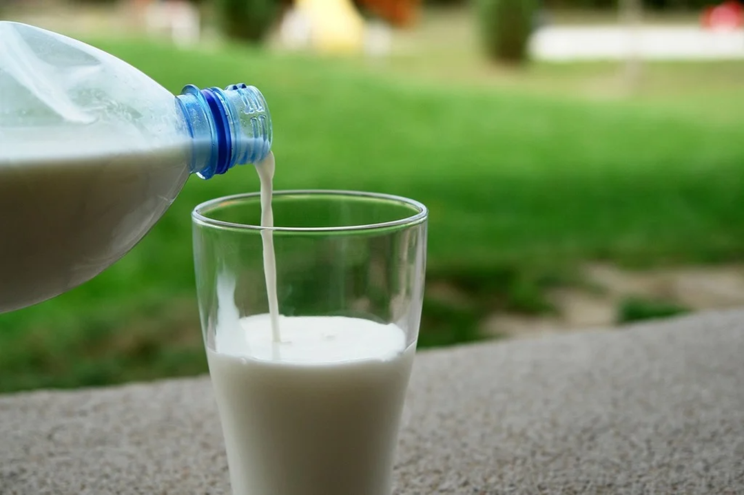 A plastic bottle pours milk into a clear glass on a stone surface, with blurred green grass and outdoor scenery in the background.