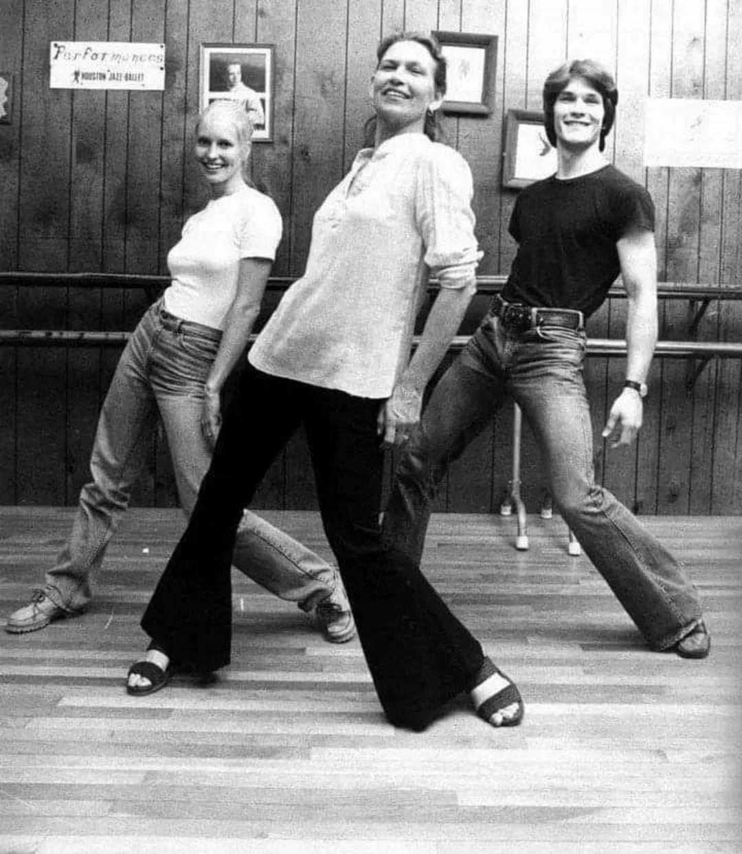 Three people pose and smile while dancing in a studio with wood-paneled walls and a barre. The two women and one man lean back with hands on their hips, showing relaxed and joyful expressions.